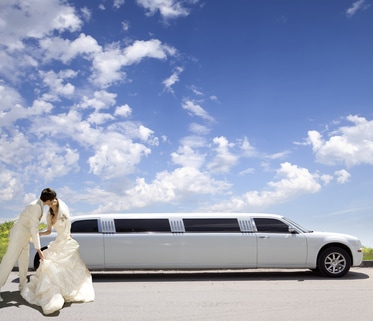 A newly married couple kisses each other while standing next to a stretch limo on a beautifully sunny day